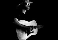 A guitarist wearing a hat plays an acoustic guitar against a dark background.