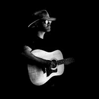 A guitarist wearing a hat plays an acoustic guitar against a dark background.
