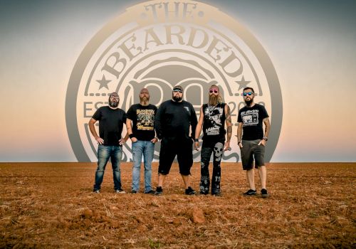 A metal band stands on a desert floor in front of a large circular logo that reads &ldquo;BEARD&hellip;,&rdquo; with five men wearing dark shirts and jeans, posing for a photo.