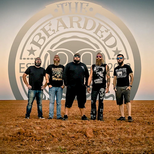 A metal band stands on a desert floor in front of a large circular logo that reads &ldquo;BEARD&hellip;,&rdquo; with five men wearing dark shirts and jeans, posing for a photo.
