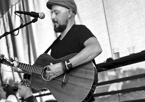 A man in a cap plays an acoustic guitar on stage, singing into a mic; another guitarist sits in the background under a shaded tent.
