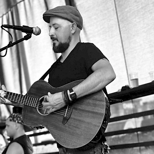 A man in a cap plays an acoustic guitar on stage, singing into a mic; another guitarist sits in the background under a shaded tent.