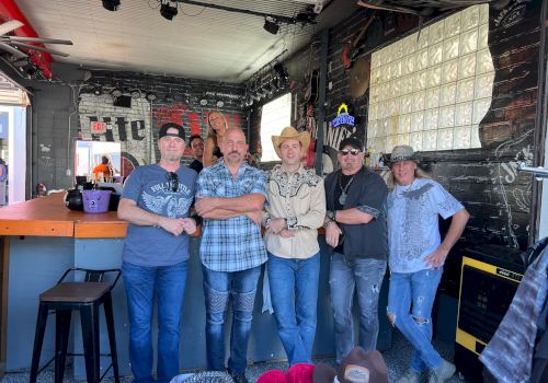 A group of six men posing together inside a rustic venue, casual attire, some hats, stage lights above, relaxed vibe, bar stools nearby.