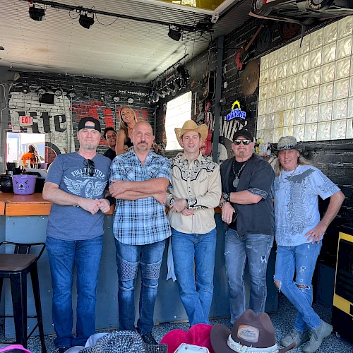 A group of six men posing together inside a rustic venue, casual attire, some hats, stage lights above, relaxed vibe, bar stools nearby.