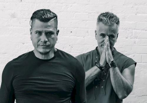 Four men posing against a white brick wall; casual, stylish, serious expressions, one with hands in prayer.