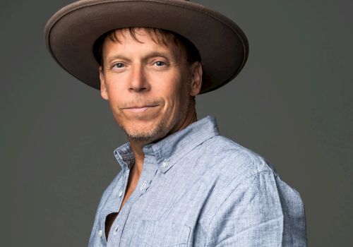 A man wearing a light blue shirt and a wide-brimmed hat poses confidently, showing a tattooed forearm and a slight smile.