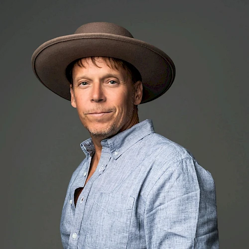 A man wearing a light blue shirt and a wide-brimmed hat poses confidently, showing a tattooed forearm and a slight smile.