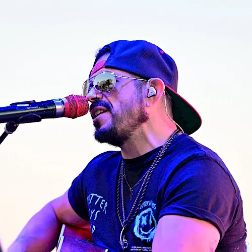 A bearded rapper in a cap and sunglasses performs into a microphone while playing an acoustic guitar onstage.