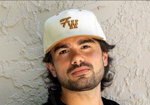 A man wearing a white cap and black shirt poses outdoors against a light wall.