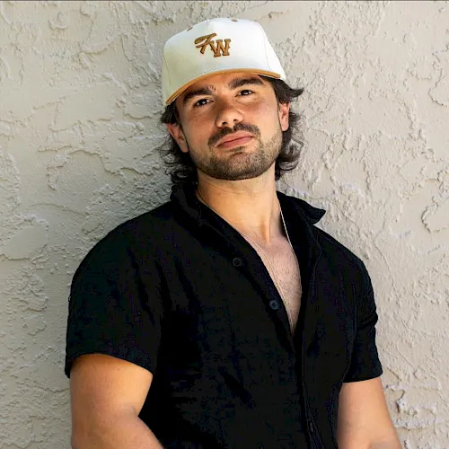 A man wearing a white cap and black shirt poses outdoors against a light wall.