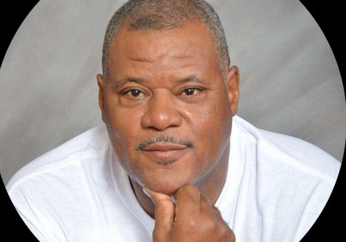 A close-up headshot of a man resting his chin on his hand against a plain background.