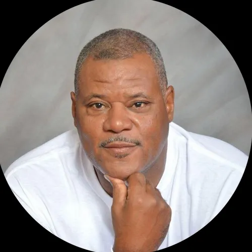 A close-up headshot of a man resting his chin on his hand against a plain background.