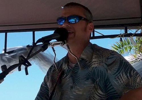 A man wearing sunglasses sings into a microphone while playing an acoustic guitar at an outdoor stage, surrounded by palm views.