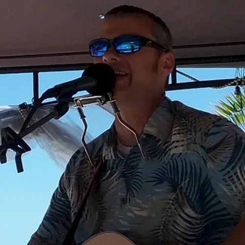 A man wearing sunglasses sings into a microphone while playing an acoustic guitar at an outdoor stage, surrounded by palm views.