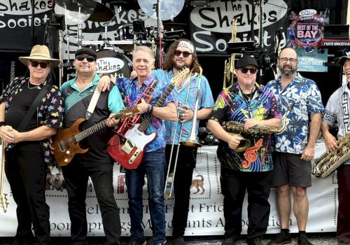 A lively band of men in colorful shirts performs outdoors on a stage, holding guitars, a saxophone, and other horns.