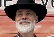 A bearded man in a black cowboy hat and patterned shirt stands outdoors in front of a red vehicle, smiling.