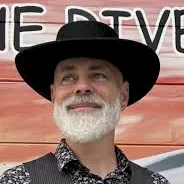 A bearded man in a black cowboy hat and patterned shirt stands outdoors in front of a red vehicle, smiling.