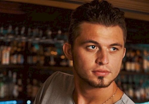 A young man with styled hair and a goatee sits in a bar, with shelves of bottles blurred in the background.