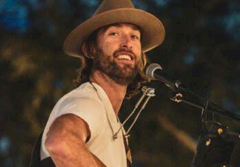 A cowboy-hat musician sings at a mic while playing an acoustic guitar on stage with drums and lights behind him.