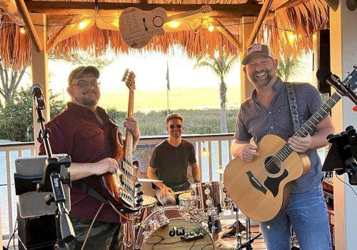 A live band plays under a tiki-style roof&mdash;guitarist and bassist up front with drums behind them at an outdoor event.