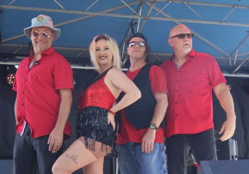 A band poses onstage under a blue tent: a blonde woman in a red sequined outfit with four men in sunglasses and red shirts.