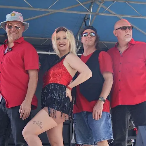 A band poses onstage under a blue tent: a blonde woman in a red sequined outfit with four men in sunglasses and red shirts.