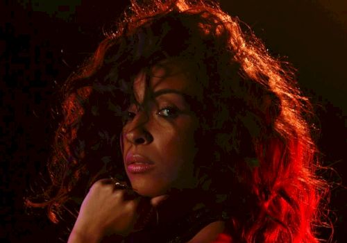 A close-up portrait of a woman with long curly hair under dramatic red-orange lighting, staring off to the side.