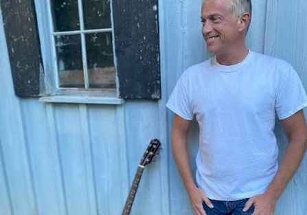 A smiling older man in a white T‑shirt stands by a blue house wall with a guitar leaning nearby.
