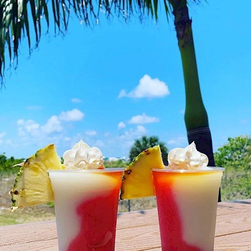 Two tropical drinks with whipped cream and pineapple slices, set outdoors under a palm tree with a clear blue sky in the background.