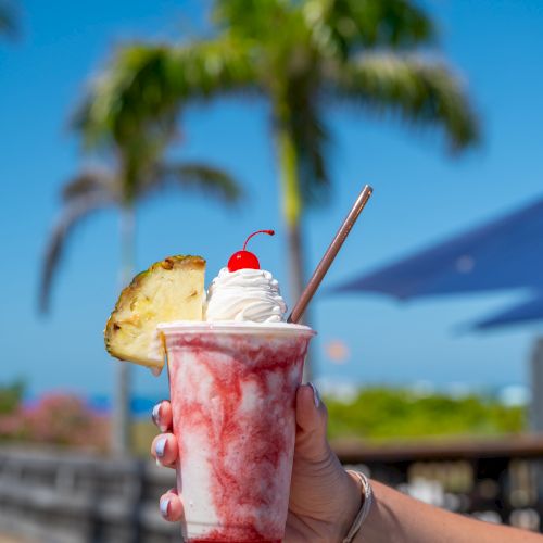 A refreshing tropical treat: a pink berry smoothie topped with whipped cream, a cherry, and a pineapple slice, served in a clear cup with a straw.