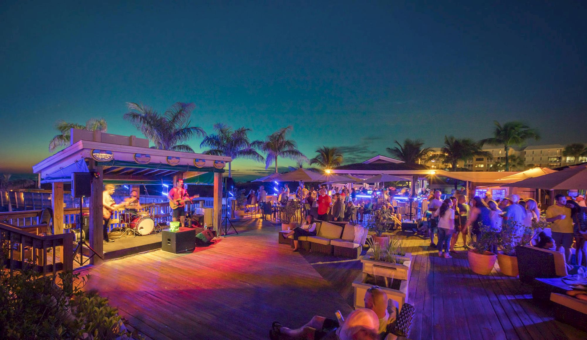 Outdoor evening event with a band performing on stage, people socializing, and vibrant lighting under palm trees and a clear sky.