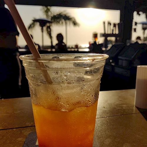 A plastic cup with an iced drink and a straw sits on a table, with a sunset and palm trees in the background. The scene looks tropical and relaxed.