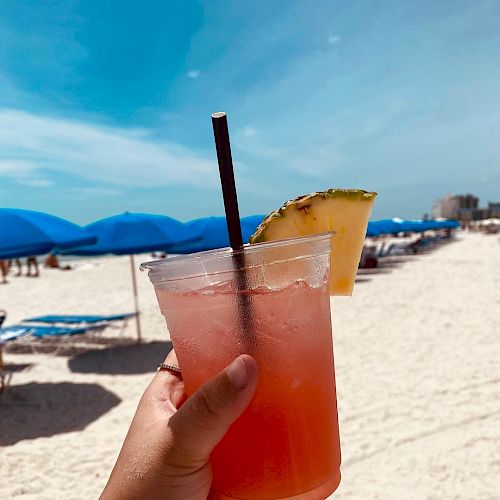 A hand holding a fruity drink garnished with pineapple against a sunny beach backdrop with blue umbrellas and lounge chairs.