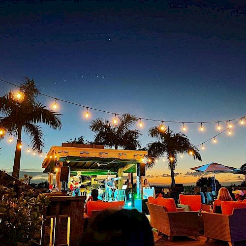 Outdoor bar setup at twilight with string lights, palm trees, seating areas, and people enjoying the ambiance and the view.