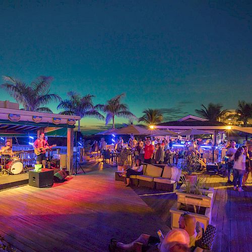 A vibrant outdoor event with live music, people socializing, and palm trees under a stunning twilight sky, creating a festive, laid-back atmosphere.