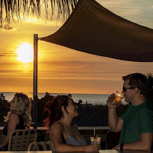 People are enjoying drinks at an outdoor bar during sunset by the beach, with a canopy overhead.