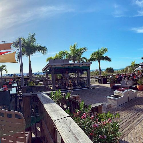 An outdoor beachside restaurant with wooden decking, palm trees, seating areas, plants, and a sunny sky in the background.