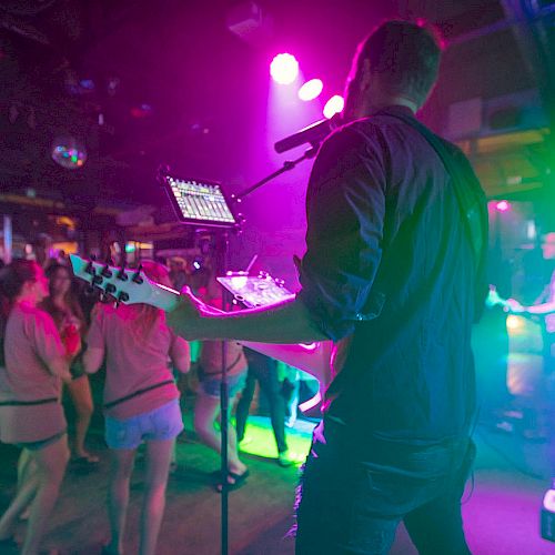 A musician playing guitar on stage is seen from behind, with a colorful, vibrant crowd enjoying the live music under dynamic lighting.