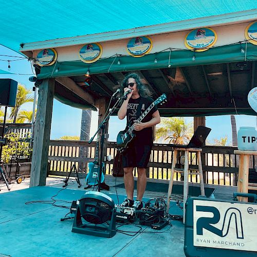 A musician stands on an outdoor stage playing a guitar and singing, with a tips jar nearby and equipment around, under a blue canopy.