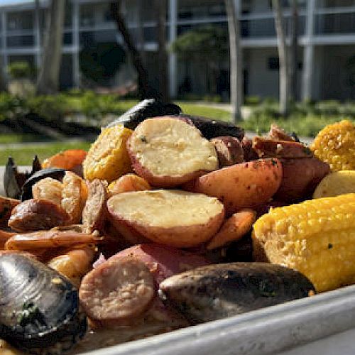 A tray filled with seafood, corn on the cob, and potatoes sits outdoors near a building with large windows.